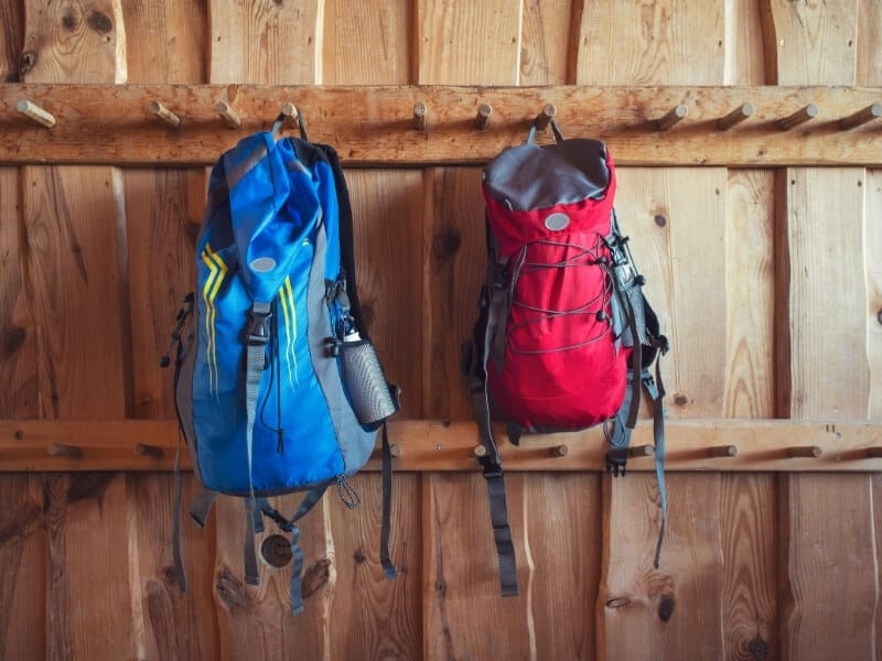 A wood paneled wall has two rows of wooden rungs hanging horizontally. On two of the rungs there are backpacks hanging. On the left is a blue backpack and on the right is a red backpack.