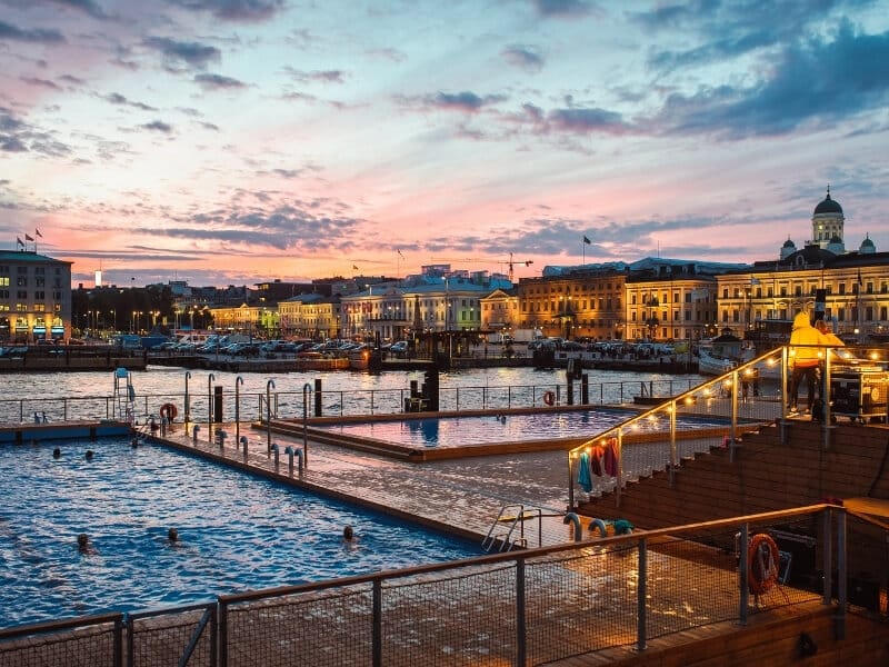 Helsinki's skyline at dusk with a pink and blue cloudy sky overhead. In the foreground is Allas Seapool, which several pools and traditional Finnish saunas in the heart of Helsinki. Photo Credit: Allas Seapool Helsinki Finland