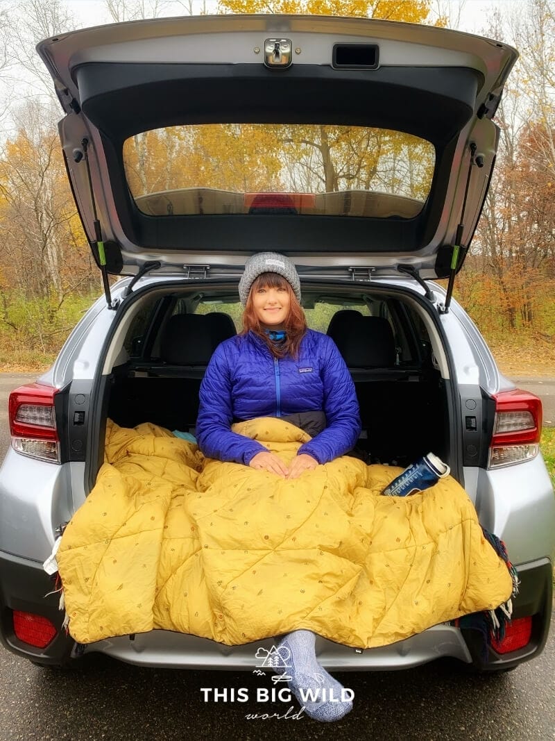 A woman sitting in the back of an SUV wearing a bright blue jacket, gray winter hat, blue wool socks covered in a yellow fluffy blanket. A blue water bottle sits on the blanket.