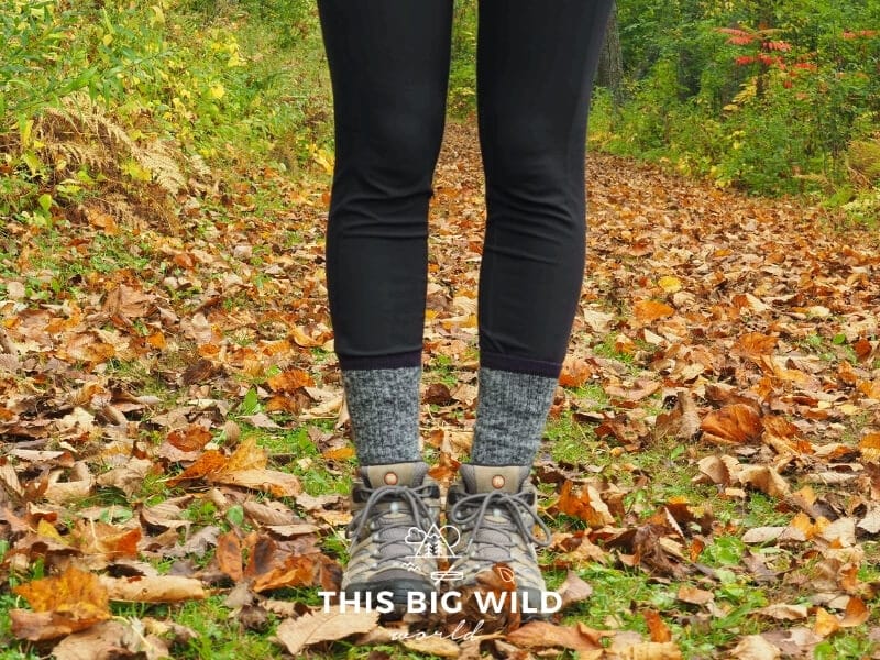 Close up of a woman's legs wearing balck leggings, black and white wool socks, and hiking boots standing in grass covered in bright red and orange leaves with grass in the background.