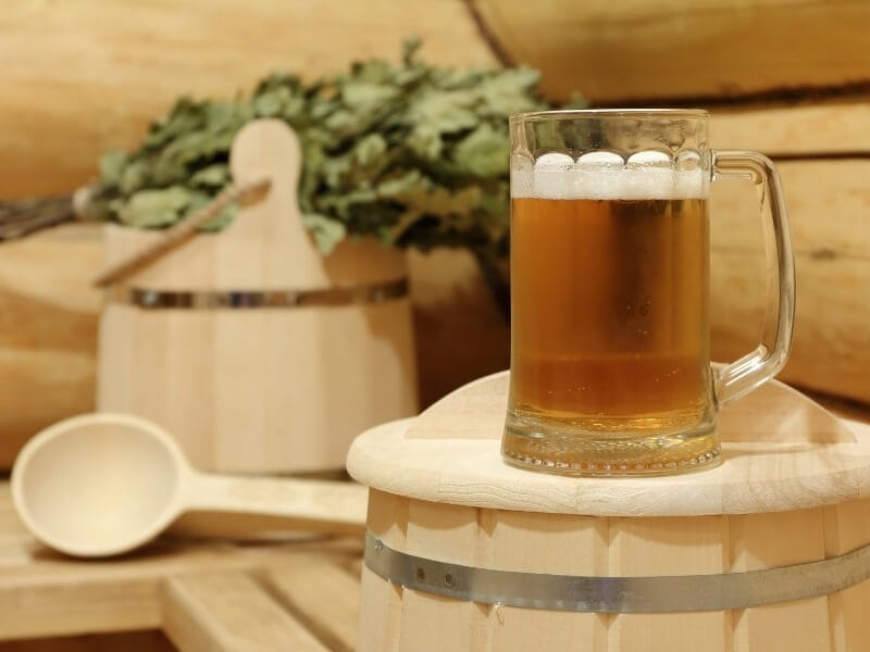 A glass of beer sits on top of a wooden barrel inside of a wood-lined sauna. In the background is a wooden bucket, spoon and birch leaves are resting on the sauna bench.