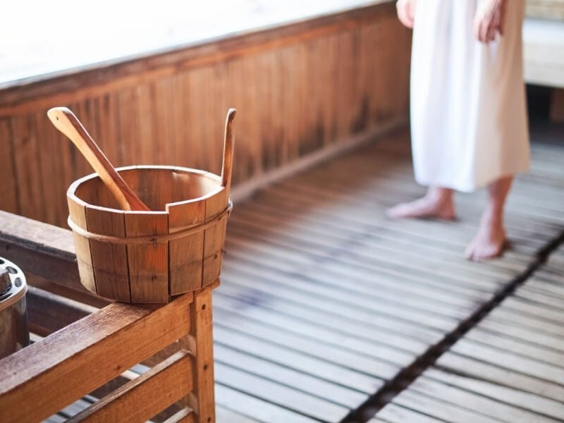 A wooden bucket with a wooden spoon rests on the edge of a railing next to the hot stones inside of a wooden sauna room.