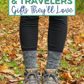 Text: Outdoor Lovers & Travelers - Gifts They'll Love Image: A woman with black leggings and black and white socks in hiking boots stands among fallen autumn leaves.
