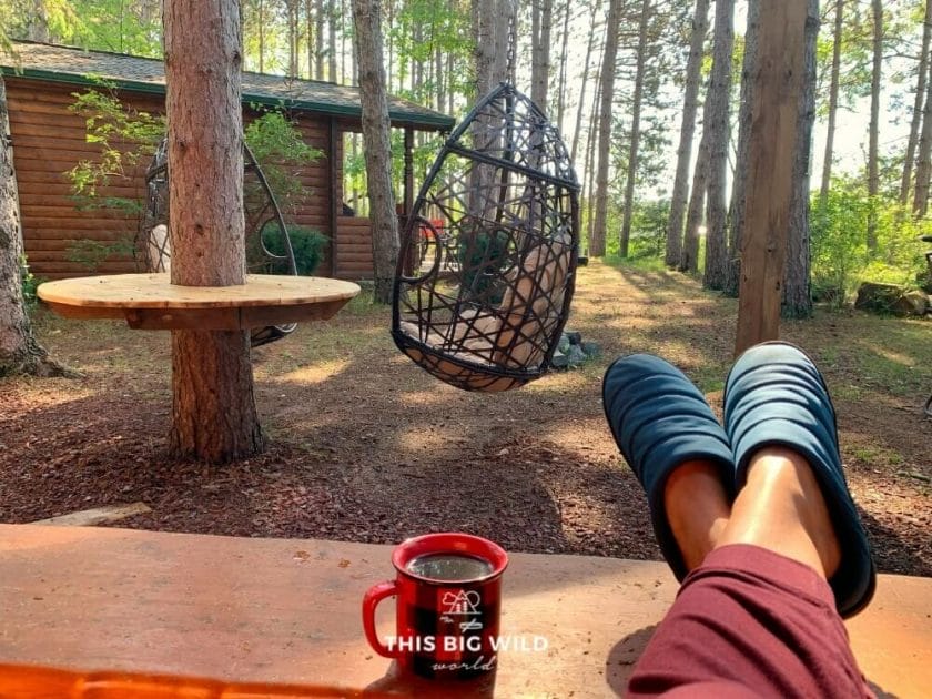 Lounging with my feet up wearing a pair of camp sandals in front of a cabin with a cup of coffee.