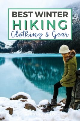 Text: Best winter hiking clothing and gear Image: Woman standing in snow leaning against a rock at the end of a calm blue lake. In the distance are snow covered mountains. 