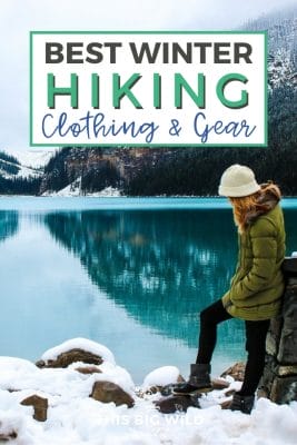 Text: Best winter hiking clothing and gear Image: Woman standing in snow leaning against a rock at the end of a calm blue lake. In the distance are snow covered mountains.
