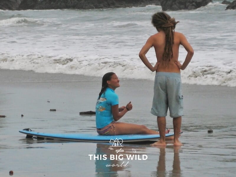 Me in a turquoise rash guard and bathing suit bottoms sitting on a surfboard on the beach. A man with long dreadlocks stands next to me encouraging me to keep trying. Behind us the waves crash on a stormy gray day near Jaco, Costa Rica.