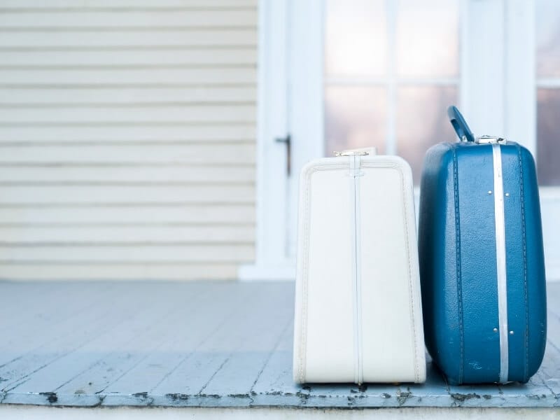 Two old-fashioned suitcases sit side by side on a wooden porch in front of a white house.