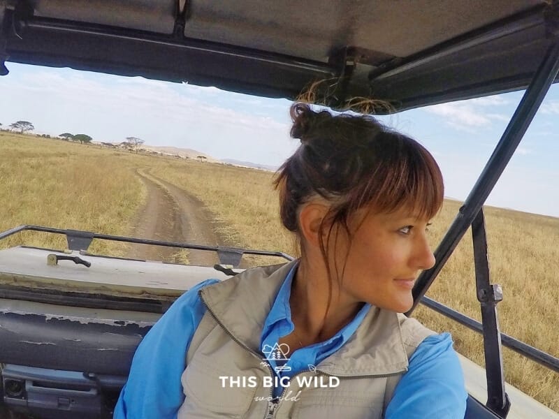 Me, a woman, with a messy bun looks to the right out of the frame. I am standing in a safari vehicle with the top raised and a dirt road with tall yellow grass behind us to the horizon. 