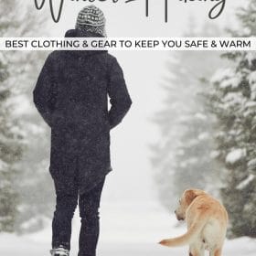 Text: Winter Hiking - Best clothing and gear to keep you safe and warm Image: Person walking away from the camera with a yellow lab walking alongside them on a snowy trail lined with pine trees.