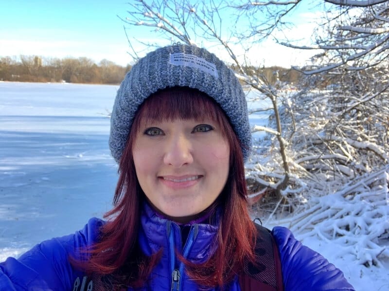 Me with red hair wearing a gray beanie and blue winter jacket hiking along a frozen snow-covered lake in Minneapolis, Minnesota. 