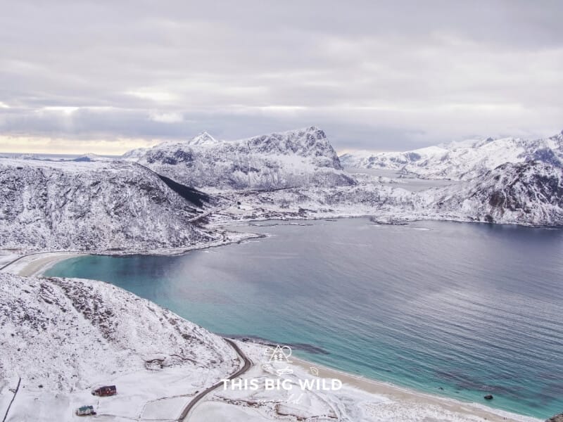 Snow covers the dark rock jutting out of the fjords in the Lofoten Islands in Norway. The water is bright blue as seen from above on the hike to Mt Mannen.