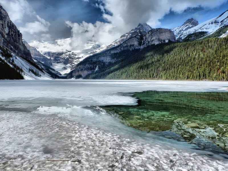 The bright blue green water of Lake Louise peeks through a coating of ice at the base of the snow-covered Canadian Rockies in Banff National Park.