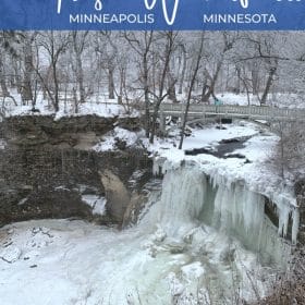 Text: Frozen Waterfalls Minneapolis Minnesota Image: Water flows under a bridge and down a waterfall that is mostly frozen creating a large icicle into a frozen pool. Surrounding the waterfall is a forest covered in a perfect dusting of snow.