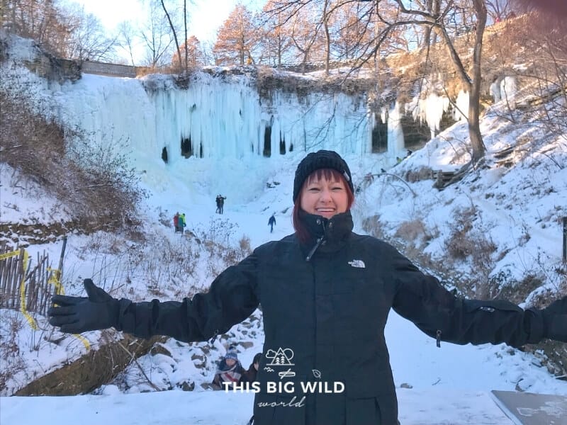 In the foreground, I am bundled up in black winter clothes with just my face showing. Behind me is a completely frozen Minnehaha Falls in Minneapolis.