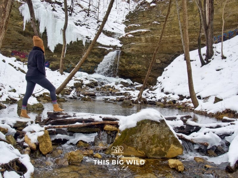 A partially frozen waterfall drops down into a stream, surrounded by limestone rock formations lined with snow and icicles. On the left, I'm standing in black with a tan hat and boots looking away from the camera at the waterfall.