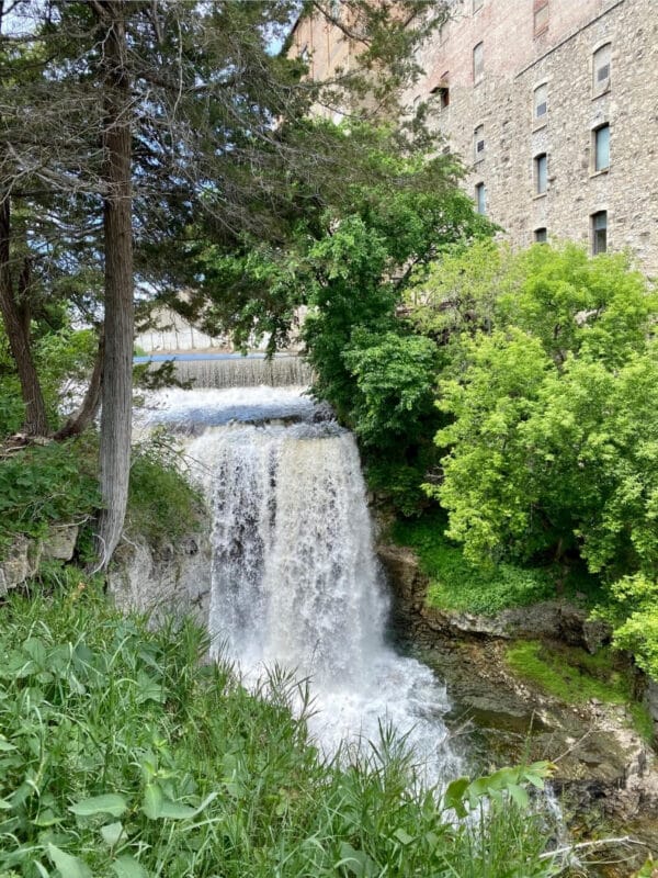 Vermillion Falls flowing rapidly along an old brick building in downtown Hastings Minnesota. Both sides are lines with lush green trees and shrubs. Photo Credit: Expedition Kristen