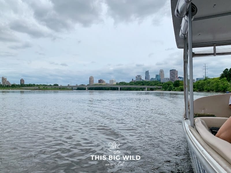A partial view of the Minneapolis skyline is visible from the Minneapolis Water Taxi as it glides through the Mississippi River on a cloudy day.