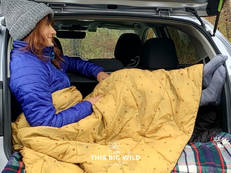 Me sitting in the back of my vehicle while car camping in the fall. Wearing a gray hat, blue jacket and a golden Rumpl Nanoloft blanket over my legs.
