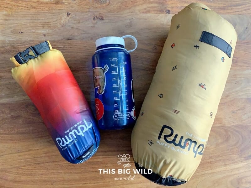 A travel size Rumpl Nanoloft blanket in a rainbow design is sitting next to a Nalgene bottle for size comparison on a wooden surface. Next to it is a larger 1-person size Rumpl Nanoloft blanket in a golden design.