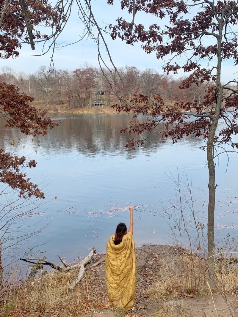Me standing on the edge of a lake in fall wearing a golden Rumpl Nanoloft blanket as a cape with one fist raised in the air.