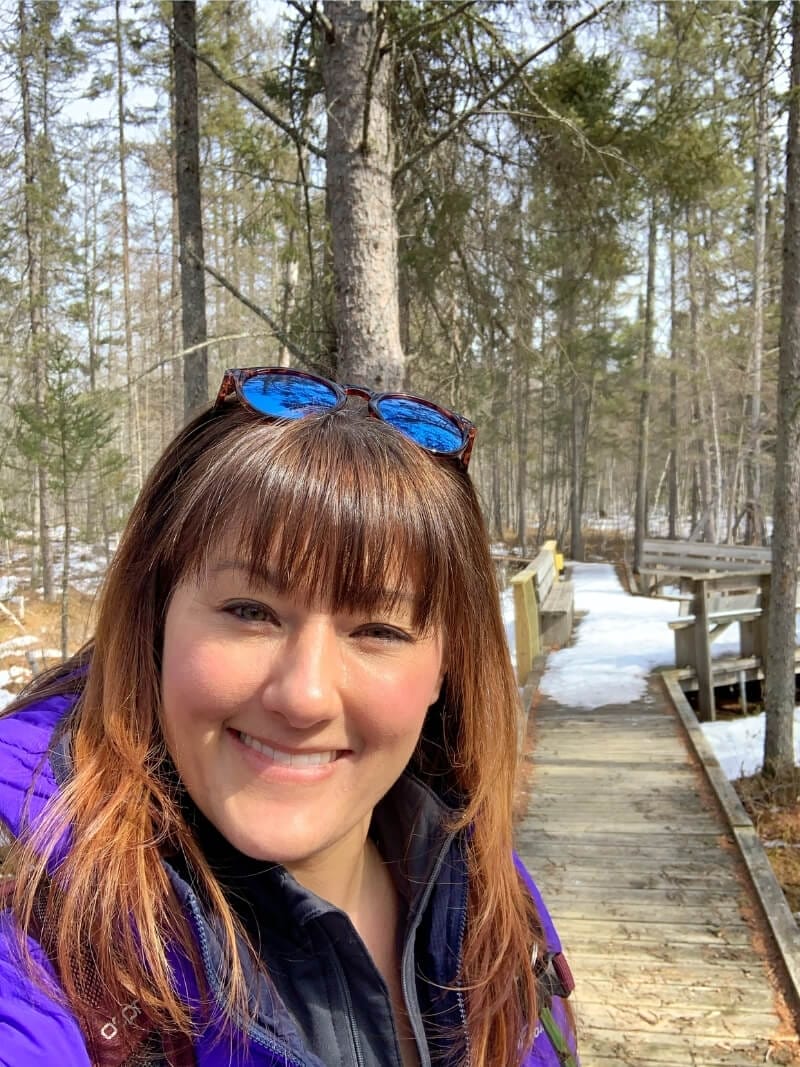 A close up of me with a smile on a warm sunny day at the end of winter in Mille Lacs Kathio State Park in Minnesota. Behind me is a boardwalk over a bog in the forest, covered in snow.