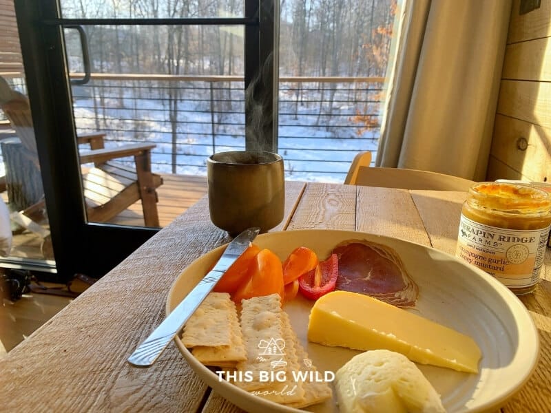 A charcuterie and cheese plate with locally sourced mustard and a steaming cup of coffee on a wood table looking out onto the patio at Cuyuna Cove.