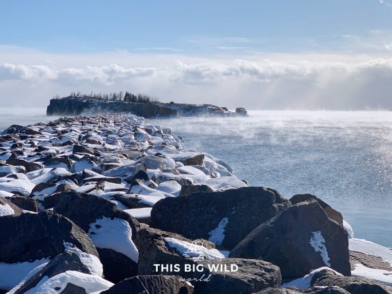 Snow covered rocks stretch out into Lake Superior towards Pellet Island on a sunny winter day. A low fog hangs over the water.