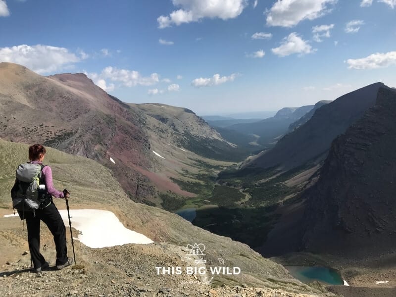Me wearing a large daypack, long sleeve top and black pants carrying trekking poles enjoying the view from the pass on the Siyeh Pass Trail at Glacier National Park.