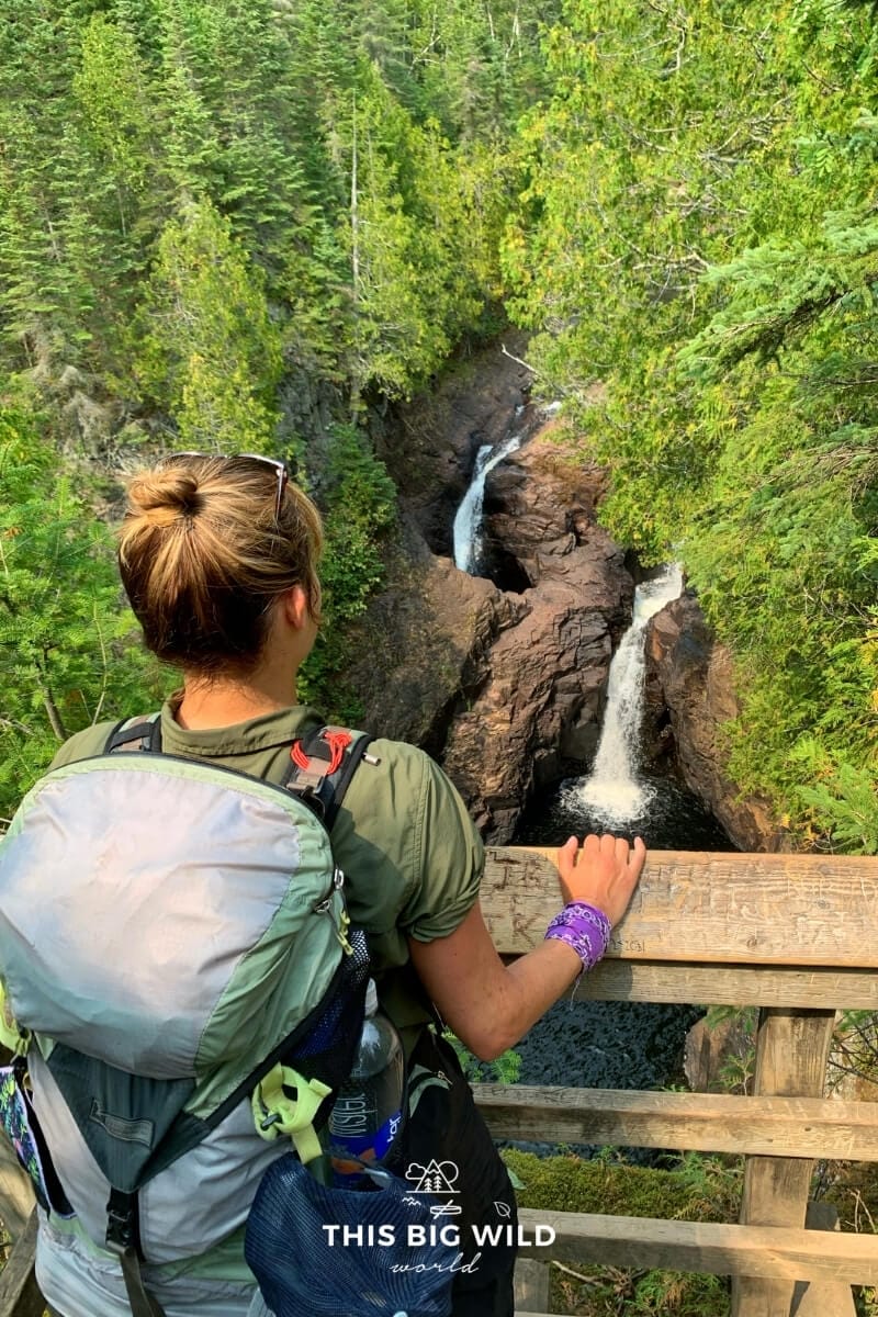 A woman is wearing a backpack while looking over a wooden railing towards a double waterfall along the Superior Hiking Trail.