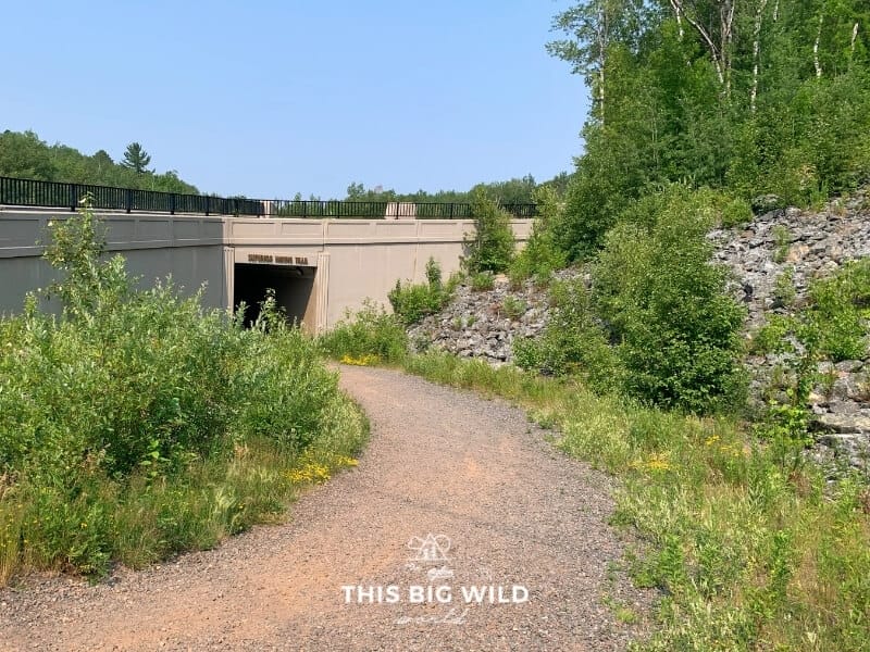 A gravel path leads downhill to a tunnel labeled 'Superior Hiking Trail', which is the entrance to the Brewer Park Loop Trail in Duluth.