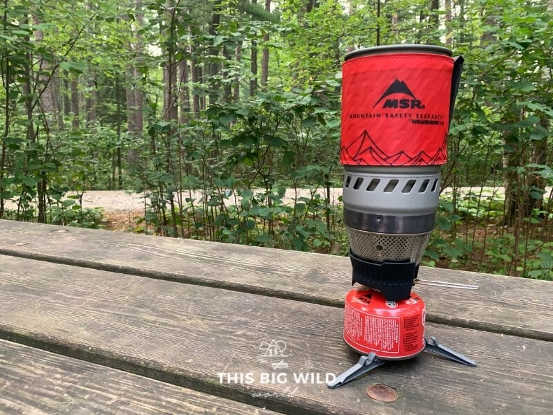 The MSR Windburner backpacking stove sits on a wooden table fully assembled with a red insulated container and wind shield.