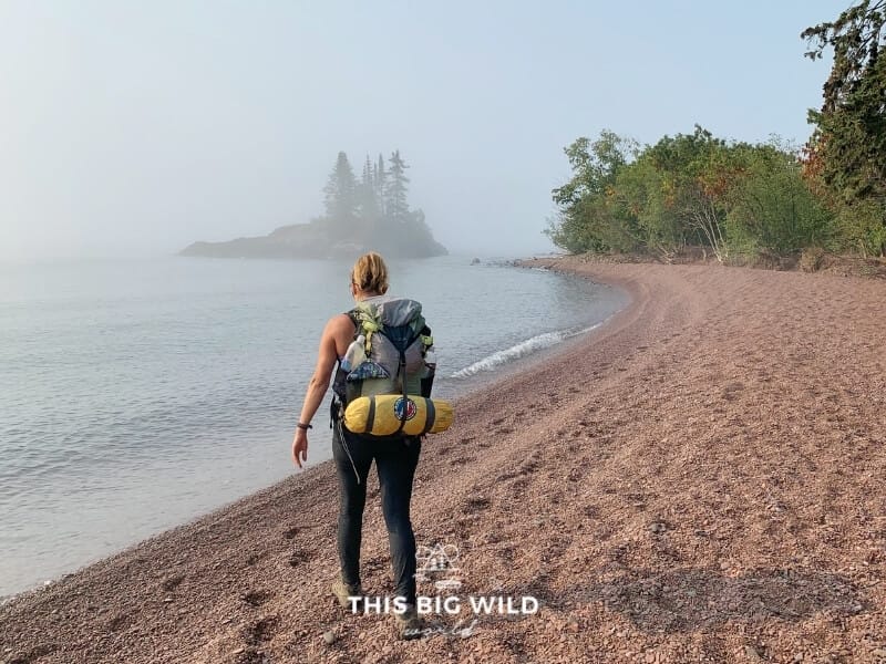 A woman walks away from the camera along Lake Superior on a foggy morning, carrying a backpack with a tent strapped securely at the bottom of it.