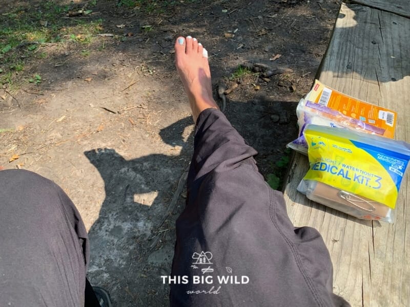 A woman's foot is hanging off of a wooden bench next to a first aid kit and a package of moleskin which is used for blisters.