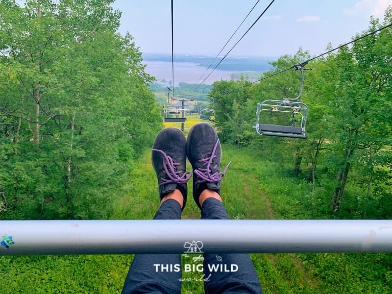 My feet stretched out in front of me while riding the scenic chair lift at Spirit Mountain in Duluth. The St Louis Bay is in front of me and green forest is on either side.
