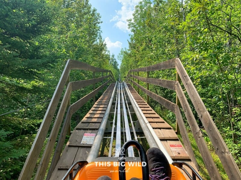 View from inside the Timber Twister alpine coaster shows my feet resting in front of me and a long wooden and metal track extending up in front of me as I get towed back up to the top.