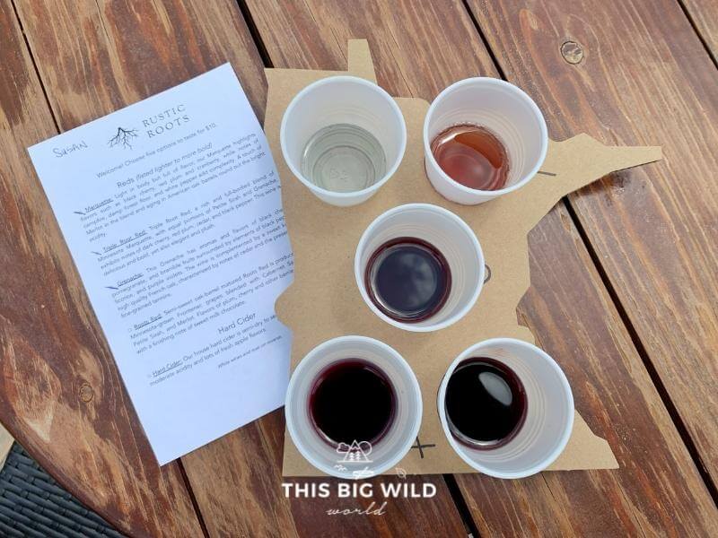 A wine tasting menu for Rustic Roots winery next to a cardboard cutout of Minnesota to carry your wine tasting glasses in while on their outdoor patio.
