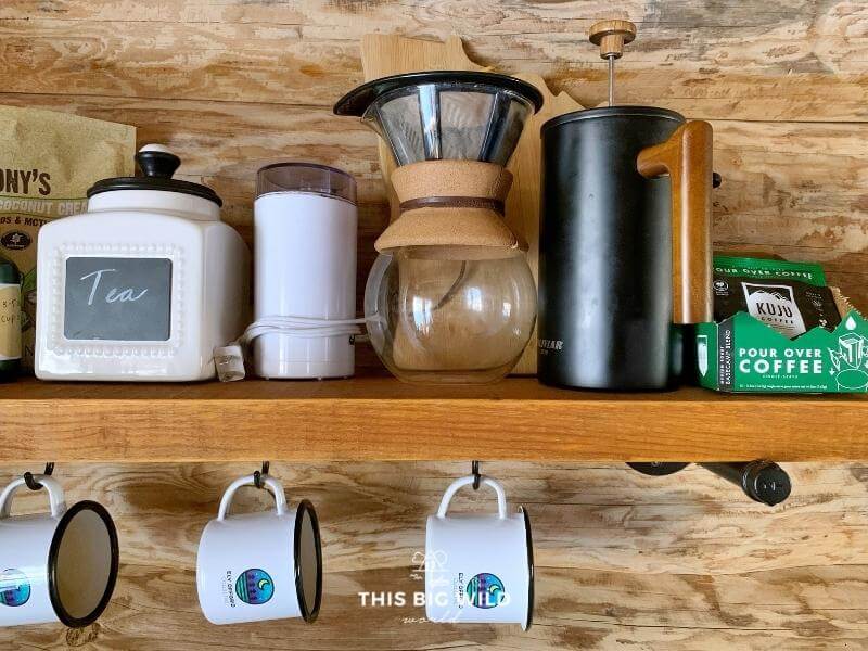 Pourover, french press coffee, and tea on a wooden shelf with coffee mugs hanging underneath.