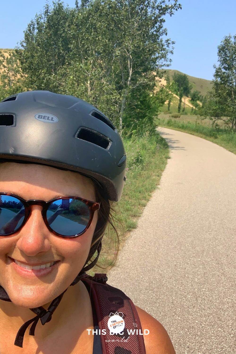 Me wearing sunglasses and a bike helmet with the Sleeping Bear Heritage Trail and sand dunes behind me.