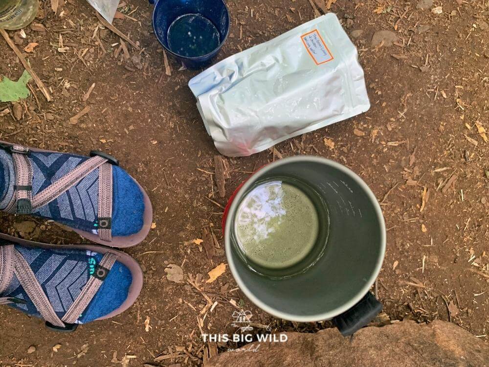Overhead shot of my feet with socks and sandals, a dehydrated meal pouch and backpacking stove on the ground.