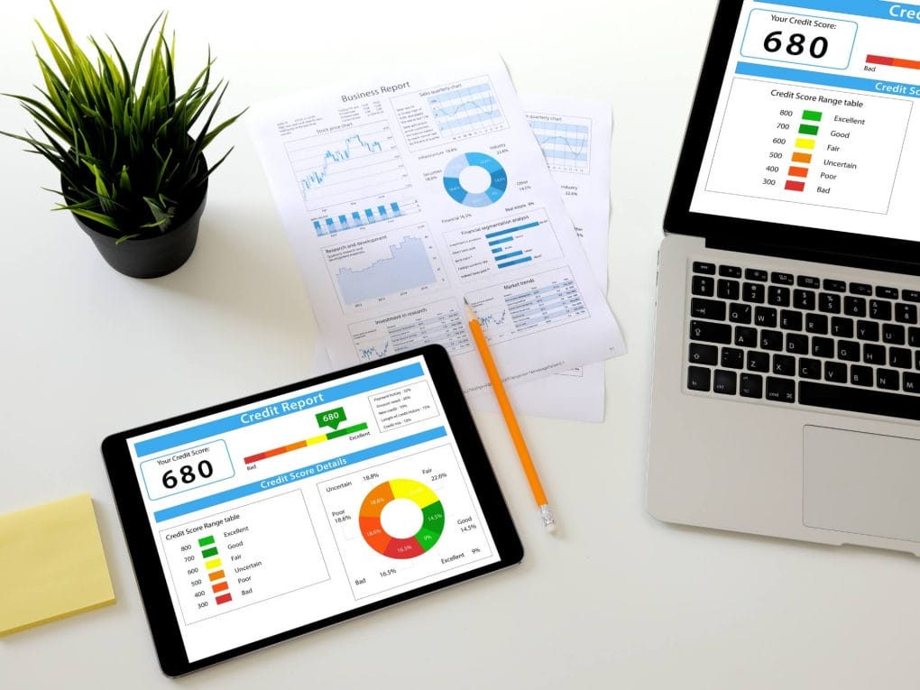 Computer and documents with credit score visible laying on table next to plant.