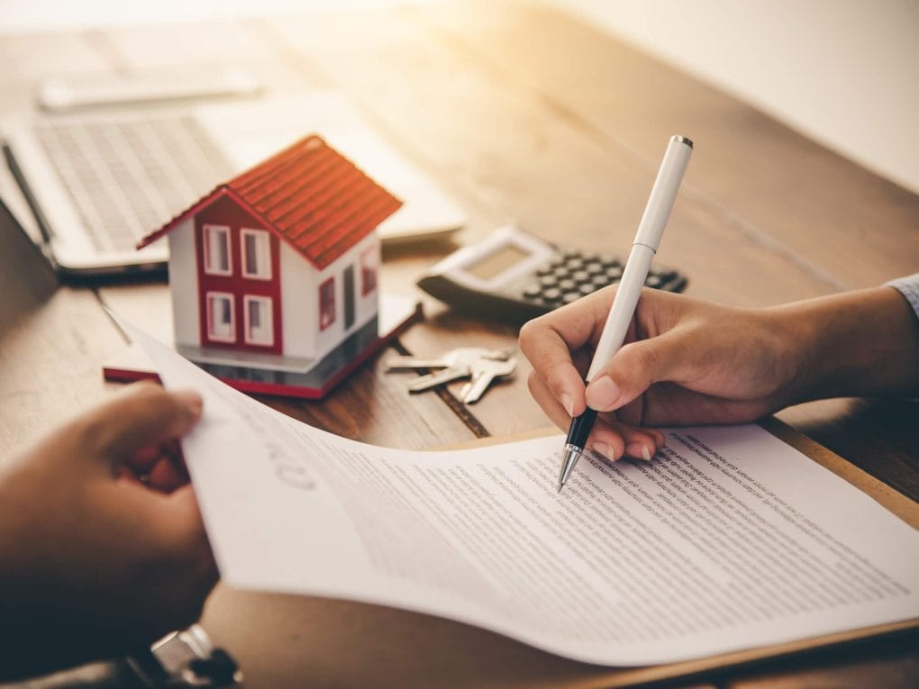 Paper being signed on table with small model of a home and calculator next to it.