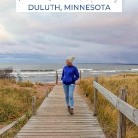 Text: Perfect Weekend Getaway Duluth Minnesota Image: Me walking on a boardwalk in front of Lake Superior with a winter hat on.
