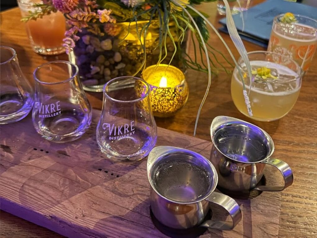 A flight of spirits with optional mixers on a wooden surface with a fresh plant in the background.