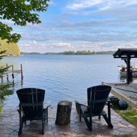 Two adirondack chairs sitting next to a lake on a sunny day.